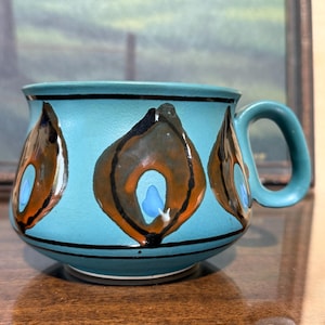 May include: A turquoise ceramic mug with a rounded body and a handle. The mug features a black rim and base, with three abstract flame-like designs in brown, orange, and blue. The mug is likely for beverages.