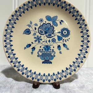 May include: Cream-colored ceramic plate with a blue floral design. The plate features a central bouquet in a vase and a border of blue floral and geometric patterns. The plate is displayed on a wooden stand.