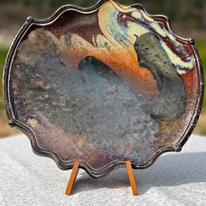 May include: A decorative ceramic plate with a scalloped edge and a dark metallic rim. The plate features a unique abstract design with shades of brown, orange, blue, and gray. It is displayed on a wooden stand, showcasing its artistic details.