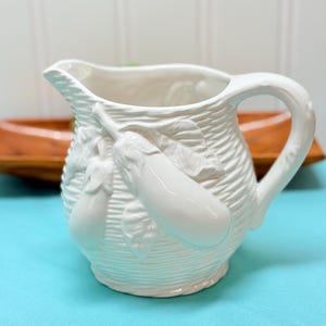 May include: White ceramic pitcher with a handle and spout. The pitcher features a textured basket weave design and is adorned with embossed eggplants and leaves. The pitcher is set against a light blue surface.