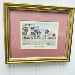 May include: A framed print featuring a coastal scene with buildings, palm trees, and a sailboat. The print is set within a gold-colored frame and a pink mat. The text "Along The Battery" is visible.