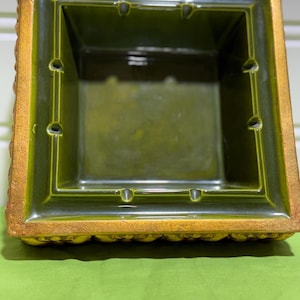 May include: A square, ceramic ashtray with a green interior and a gold-colored, textured border. The ashtray has four indentations for cigarettes and a stepped design. The exterior border has a decorative, raised pattern.