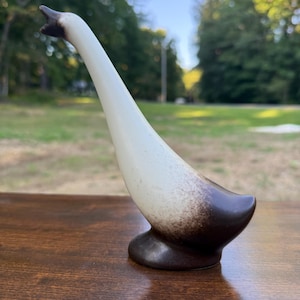 May include: A ceramic goose figurine featuring a long, slender neck and a rounded base. The body transitions from a dark brown color at the bottom and tail to a creamy off-white shade along the neck and head, with a dark brown beak.
