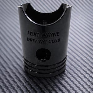 May include: Black cylindrical object with the text "FORT WAYNE DRIVING CLUB". The object has a curved top and a circular hole. The object has horizontal lines around the middle.