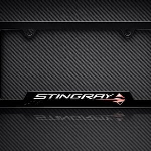 Può includere: Cornice nera per targa con la scritta "STINGRAY" in lettere bianche e un design a freccia rossa. La cornice è rettangolare e ha uno sfondo in fibra di carbonio.