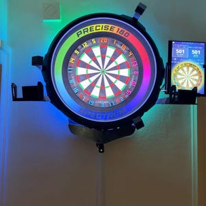 Puede incluir: Un tablero de dardos electrónico montado en la pared con un diseño circular. El tablero de dardos presenta un área de destino colorida con números y el texto "PRECISE 180". Una pantalla más pequeña muestra la puntuación. La tabla está iluminada con luces azules y verdes.