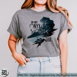 May include: A gray t-shirt with a graphic design of a dark gray storm cloud with lightning bolts and a black dragon. The text "In My Onyx Storm Era" is printed on the shirt.