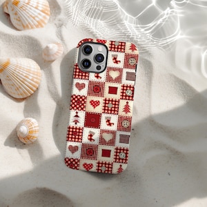 Op de afbeelding: Een rood-witte patchwork telefoonhoes met harten, knopen, kerstbomen en sokken. De hoes ligt op een zanderige ondergrond met schelpen.