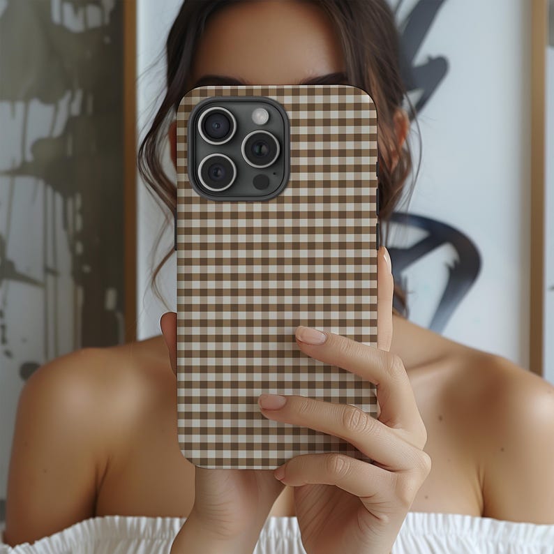 May include: A brown and white gingham patterned phone case. The case is held in front of a person's face, obscuring the features. The phone case has a square camera module with three lenses.