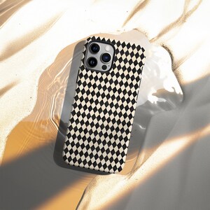 May include: A smartphone case with a black and cream diamond pattern. The case has a camera cutout and is resting on a sandy surface. The case is designed to protect the phone while adding a stylish look.