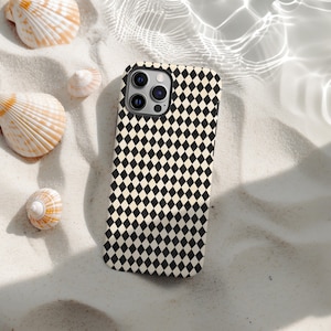 May include: A black and white diamond pattern phone case. The case is lying on white sand with seashells scattered around it. The phone case has a camera bump with three lenses.