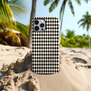 May include: A smartphone case with a black and cream diamond pattern. The case is shown on a sandy surface with palm trees in the background. The case has a camera and flash module.
