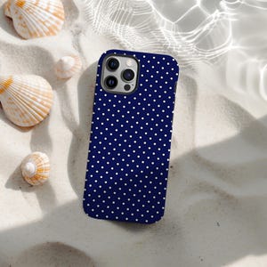 Puede incluir: Funda de teléfono azul marino con lunares blancos. La funda está sobre arena blanca, con conchas marinas cerca. La funda tiene una cámara de triple lente. El fondo tiene un efecto de agua.