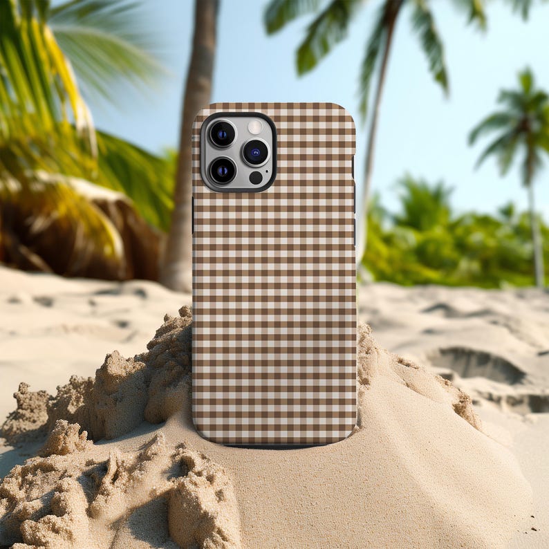 May include: A brown and white checkered phone case. The case is on a sandy surface with palm trees in the background. The phone case has a camera and flash.