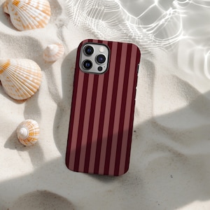 Puede incluir: Funda de teléfono a rayas verticales granate y rojo claro, con cámara de objetivos múltiples. Descansa sobre arena blanca, con conchas. Efecto de agua en el fondo. Patrón de rayas verticales.