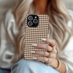 May include: A brown and white gingham pattern phone case. The case has a square pattern and a camera cutout. The phone case is held by a person with long nails and gold rings.
