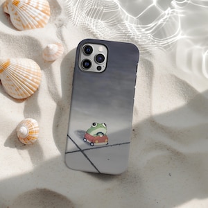 Puede incluir: Una funda de teléfono inteligente con un diseño gris degradado y una rana de dibujos animados conduciendo un coche rojo. La funda está rodeada de conchas marinas sobre una superficie arenosa.