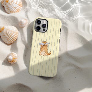 Peut inclure: Coque de téléphone jaune à rayures verticales avec une illustration à l'aquarelle d'un chat roux tenant deux poissons dans sa bouche. La coque est posée sur une surface sablonneuse avec des coquillages.