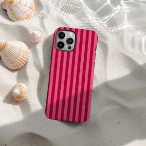 Puede incluir: Una funda de teléfono con un diseño de rayas verticales en tonos rosas y rojos. La funda está sobre arena blanca, con conchas marinas alrededor. La funda tiene un borde negro y un módulo de cámara.