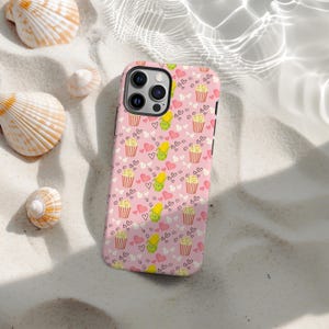 Puede incluir: Funda de teléfono rosa con un patrón repetido de cubos de palomitas de maíz, mazorcas de maíz y corazones. La funda tiene un acabado brillante y un recorte para la cámara. El fondo es una playa de arena con conchas.