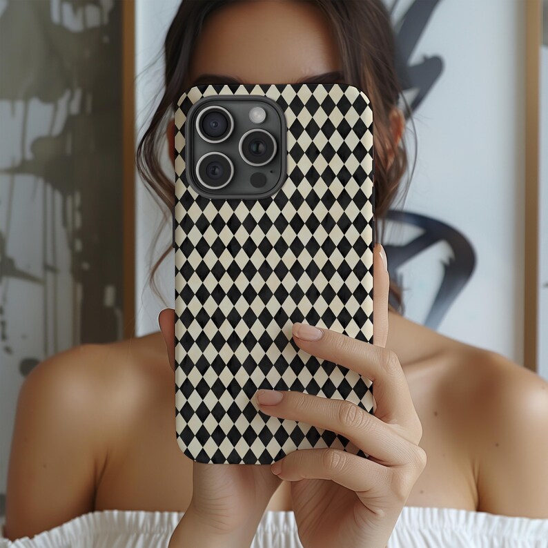 May include: A black and cream-colored diamond pattern phone case. The case has a square shape with a camera cutout on the top left. The phone case is held in front of a person's face.