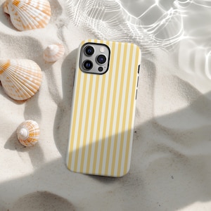May include: A smartphone case with a yellow and white striped pattern. The case is lying on a bed of sand, surrounded by seashells. The case has a black camera module.