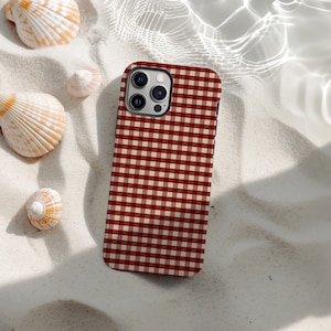 May include: A smartphone case featuring a red and cream gingham design. The case is placed on white sand, with seashells scattered around. The case has a black border and a camera module.
