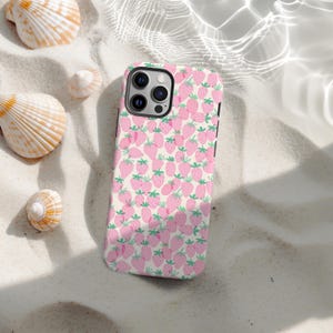 May include: A phone case with a pink strawberry pattern on a cream background. The case is lying on a sandy surface with seashells scattered around it. The strawberries have green stems and leaves.