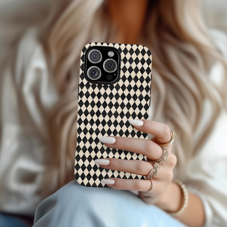 May include: A black and cream-colored diamond pattern phone case. The case has a camera cutout and is held in a hand with multiple gold rings and a bracelet. The phone case is a protective accessory.