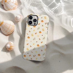 Op de afbeelding: Een crèmekleurige telefoonhoes met een patroon van kleine oranje pompoenen, bladeren en takjes. De hoes ligt op wit zand, omringd door verschillende schelpen.
