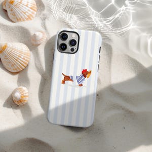 Puede incluir: Funda de teléfono con un diseño de rayas azules y blancas. La funda presenta un perro salchicha de dibujos animados con boina roja y camisa a rayas azules y blancas, sosteniendo una baguette. La funda está rodeada de conchas sobre una superficie arenosa.