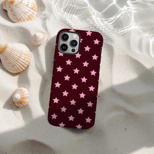 Könnte beinhalten: Eine weinrote Handyhülle mit einem Muster aus rosa Sternen. Die Hülle liegt auf weißem Sand, umgeben von Muscheln. Die Handyhülle hat ein Kameramodul mit drei Linsen. Der Hintergrund deutet auf einen Strand hin.