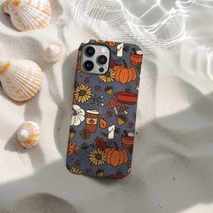 May include: A mobile phone case with a seasonal autumn design. The pattern includes pumpkins, sunflowers, acorns, and coffee cups in orange, yellow, and brown on a grey background. The case is on a sandy surface with seashells.