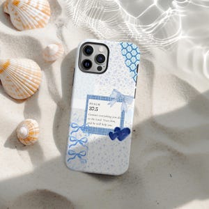 Puede incluir: Una funda de teléfono blanca y azul con un diseño floral y lazos. La funda presenta el texto "PSALM 37:5" y una cita. La funda está rodeada de conchas marinas sobre una superficie arenosa.