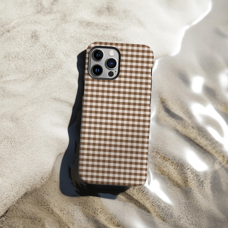 May include: A brown and white gingham patterned phone case. The case has a square pattern and a camera cutout. The phone case is resting on a sandy surface.