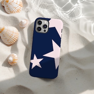 May include: A navy blue phone case with a pattern of large, light pink stars. The case is lying on a bed of white sand, surrounded by seashells. The phone case has a black camera module.