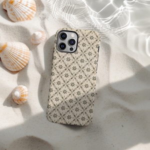 May include: A beige phone case featuring a repeating floral design in dark grey. The case is on white sand, with seashells around it. The phone case has a camera bump with three lenses.