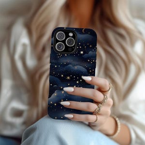 Pode incluir: Capa de telefone azul escuro com um design celestial de estrelas douradas, nuvens e uma lua crescente. A capa tem um acabamento brilhante e um recorte para a c&acirc;mera. A capa do telefone est&aacute; na m&atilde;o de uma pessoa.