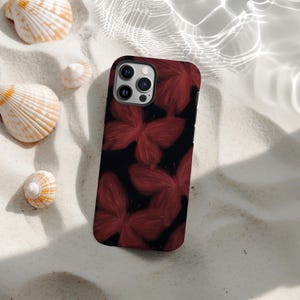 Könnte beinhalten: Eine schwarze Smartphone-Hülle mit einem Muster aus roten Schmetterlingsillustrationen. Die Hülle liegt auf einer sandigen Oberfläche, mit Muscheln in der Nähe. Die Hülle hat ein Kameramodul mit drei Objektiven.