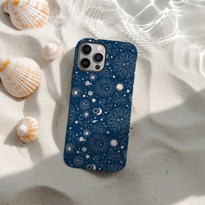 Pode incluir: Capa de telemóvel azul marinho com um design celestial com sóis, luas, estrelas e olhos. A capa está sobre areia branca, rodeada por conchas.