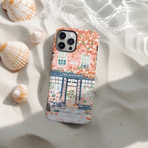May include: A phone case with a bookstore illustration, text reads "THE BOOKSHOP". The design includes a pink and orange floral pattern and a detailed shop front. The case is set on a sandy surface with seashells. The shop front has a bicycle.
