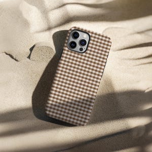 May include: A brown and white gingham patterned phone case. The case is made of a hard material and has a camera cutout. The case is resting on a sandy surface.