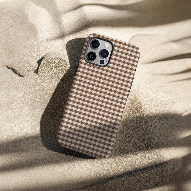 May include: A brown and white gingham patterned phone case. The case is made of a hard material and has a camera cutout. The case is resting on a sandy surface.