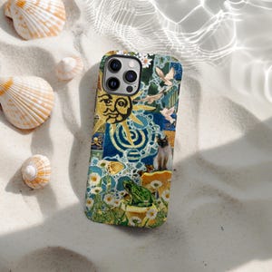 May include: A patterned phone case with a collage design. The case features a sun, a cat, a frog, and flowers, set against a blue and yellow background. The case is surrounded by seashells on a sandy surface.