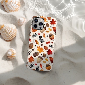 Puede incluir: Funda de teléfono inteligente con un patrón de temática otoñal con hojas, bellotas y piñas en tonos rojos, naranjas y marrones. La funda está colocada sobre una superficie arenosa con conchas marinas.