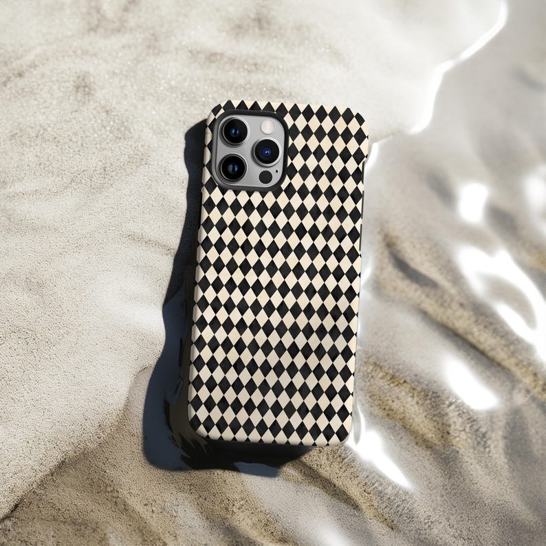 May include: A smartphone case with a black and cream diamond pattern. The case has a camera cutout and is resting on a sandy surface. The case is designed to protect the phone from scratches and damage.
