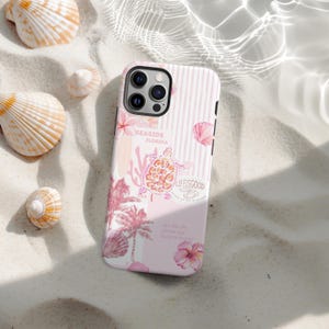 Puede incluir: Funda de teléfono a rayas rosas y blancas con un diseño de temática playera. La funda presenta una tortuga, palmeras, conchas marinas y las palabras "SEASIDE FLORIDA" y "LIFE IS GOOD". Conchas marinas rodean la funda sobre una superficie arenosa.