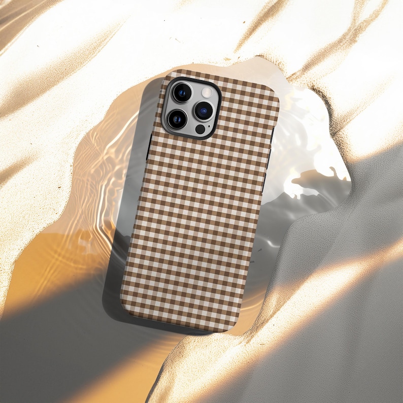 May include: A brown and white gingham pattern phone case. The case has a black border and a camera cutout. The case is lying on a textured surface with shadows.