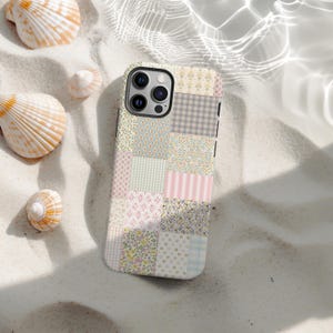 以下が含まれることがあります： パステルカラーのパッチワークデザインのスマートフォンケース。ケースには、花柄、チェック柄、ストライプ柄など、さまざまなパターンの正方形が使用されています。ケースは、貝殻のある砂浜の上に置かれています。
