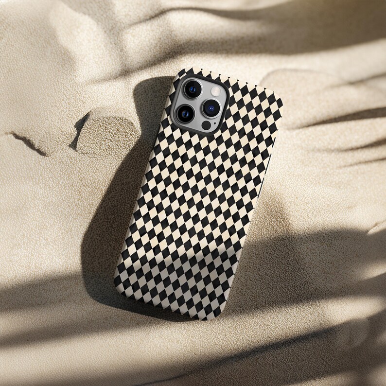 May include: A smartphone case with a black and white checkered pattern. The case is lying on a sandy surface. The case has a camera module with three lenses.